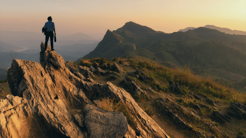 A leader standing at the top of a mountain, looking off into the distance as the sun sets.